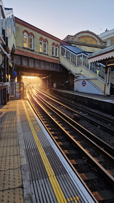 This image depicts a section of an outdoor train station platform with textured yellow tactile paving along the edge for safety. There are multiple sets of railway tracks curving into the distance, passing underneath a bridge that features a brick and metal structure with windows and a staircase leading up to an enclosed footbridge. The station is illuminated by natural light, with the sun setting on the horizon, casting a warm glow through the bridge opening. A curved wall with architectural details and a sign referencing the London Underground is visible to the right. The scene is set during daylight, with no visible moving trains or passengers, illustrating a quiet moment before scheduled train arrivals. This setting emphasizes the importance of clear, safe pathways for passengers during home relocation or furniture transport arrived in train stations, with [COMPANY_NAME] often involved in assisting with moving logistics and transportation services near Finsbury Park.
