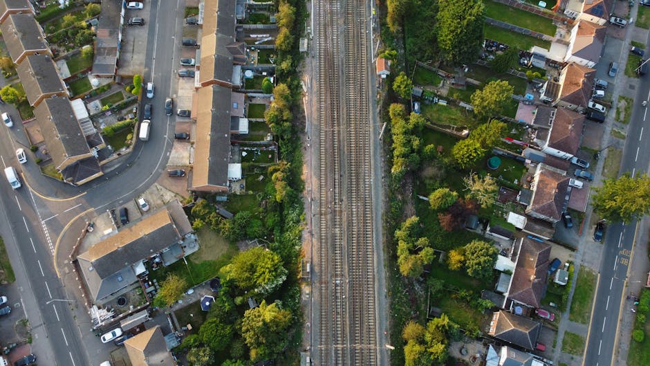 An aerial view showing a residential neighbourhood with detached and semi-detached houses, some with back gardens and trees, adjacent to railway tracks running vertically through the centre of the image. The houses have tiled roofs, and streets with parked cars border the properties. A section of a road with a curved junction and multiple parked vehicles is visible on the left, while another street runs parallel to the railway on the right. The image captures the scene during daytime with natural lighting, providing a clear view of the layout and environment typical of a home relocation area. This setting aligns with home removals and furniture transport services offered by [COMPANY_NAME], illustrating a typical urban environment where moving logistics are managed and executed.