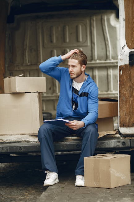 A man sitting on the edge of an open moving van, surrounded by cardboard boxes of various sizes, some sealed with packing tape and others open with packing material visible. He is wearing a blue hoodi