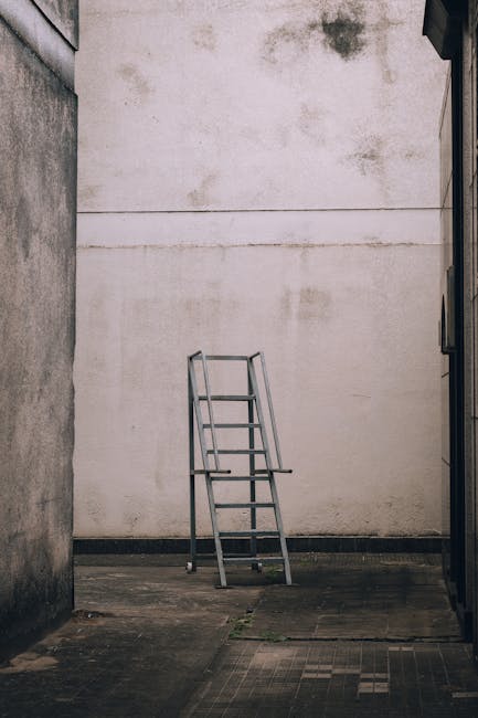 A metal step ladder with four steps and safety rails is positioned outdoors on a paved surface between two narrow building alleyways, leading towards a large white wall with visible weathering and stains. The area appears to be part of a residential or commercial exterior space, possibly used for access or maintenance purposes. This scene, captured in natural light, reflects the initial stage of a home relocation or moving process, where equipment like ladders may be prepared for furniture or appliance transport. The image is relevant to house removals services provided by [COMPANY_NAME], illustrating typical site preparation or access points prior to loading or unloading furniture and other household items, which may involve packaging, lifting, or moving activities within or outside the property.