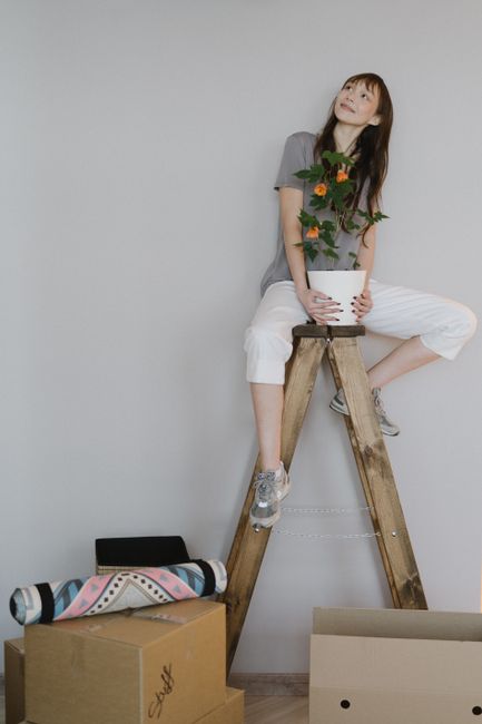 A young woman with shoulder-length brown hair and light skin sits on a wooden stool positioned against a plain, light-colored wall. She is wearing a gray t-shirt, white cropped pants, and gray sneakers, and appears to be holding a white pot with green leafy plants and orange flowers. In front of her on the floor are several cardboard boxes, one of which is partially open, along with a rolled-up fabric blanket with a geometric pattern. To the left on the floor, there is a black tray or laptop case. The scene suggests a home relocation or packing process, with the woman taking a break or posing during an interior moving or unpacking activity in a modern, minimalistic setting, related to house removals or furniture transport services provided by [COMPANY_NAME] as part of a professional move.