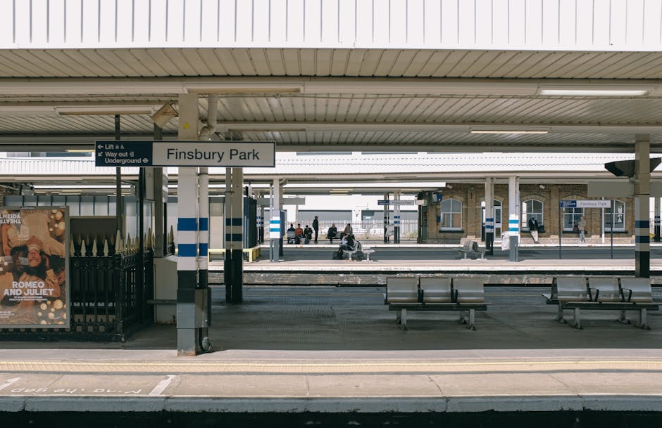 The image shows the Finsbury Park train station platform during daylight hours, with a large white sign displaying the station name 'Finsbury Park' mounted under the roof ceiling. Several metal benches are positioned along the platform for passenger seating. The platform surface features tactile paving and yellow safety lines near the edge. In the background, additional platforms are visible, with a few passengers waiting or walking, some seated on benches, and others standing or heading towards exits. To the left, there is a small advertisement poster enclosed in a glass frame. The station infrastructure includes supporting columns with blue and white striped markings, and overhead lighting fixtures are installed on the ceiling. Outside the station, the surroundings are partly visible, including a brick building with windows on the far side, and people are seen walking or sitting in the distance, capturing a typical scene of a busy urban transport hub. This scene highlights the infrastructure involved in home relocation or furniture transport, with the station serving as a key transit point for moving logistics.