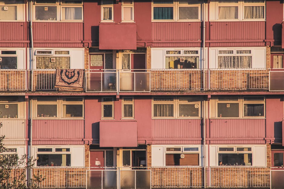 A close-up view of an apartment building exterior with multiple balconies, each enclosed with glass and metal railings. The building features pink and white paneling, with some balconies displaying personal items such as a patterned rug hanging over a railing. Several windows on different floors are visible, some with curtains drawn, and others with blinds. The scene is illuminated by natural daylight, highlighting the textures of the building materials. This image reflects typical residential flats in Finsbury Park, where house removals and furniture transport often involve navigating multi-storey layouts and coordinating packing and loading activities with professional move teams like [COMPANY_NAME], utilizing equipment such as trolleys and blankets to assist in the safe transfer of belongings during the home relocation process.