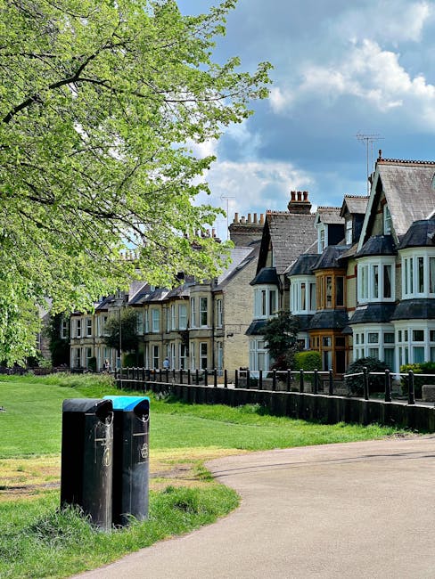 A row of traditional terraced houses with bay windows and pitched roofs, situated behind a low black metal fence along a residential street in Finsbury Park. In the foreground, there is a grassy verge with a large leafy tree on the left side, and a pair of black plastic wheelie bins placed on a concrete footpath. The bins are positioned next to each other near the edge of the grass, and the pavement extends towards the houses. The sky above is partly cloudy with patches of blue, and the scene captures a quiet urban neighbourhood environment typical for house removals or relocation logistics, supporting property-based moving services like those offered by [COMPANY_NAME].